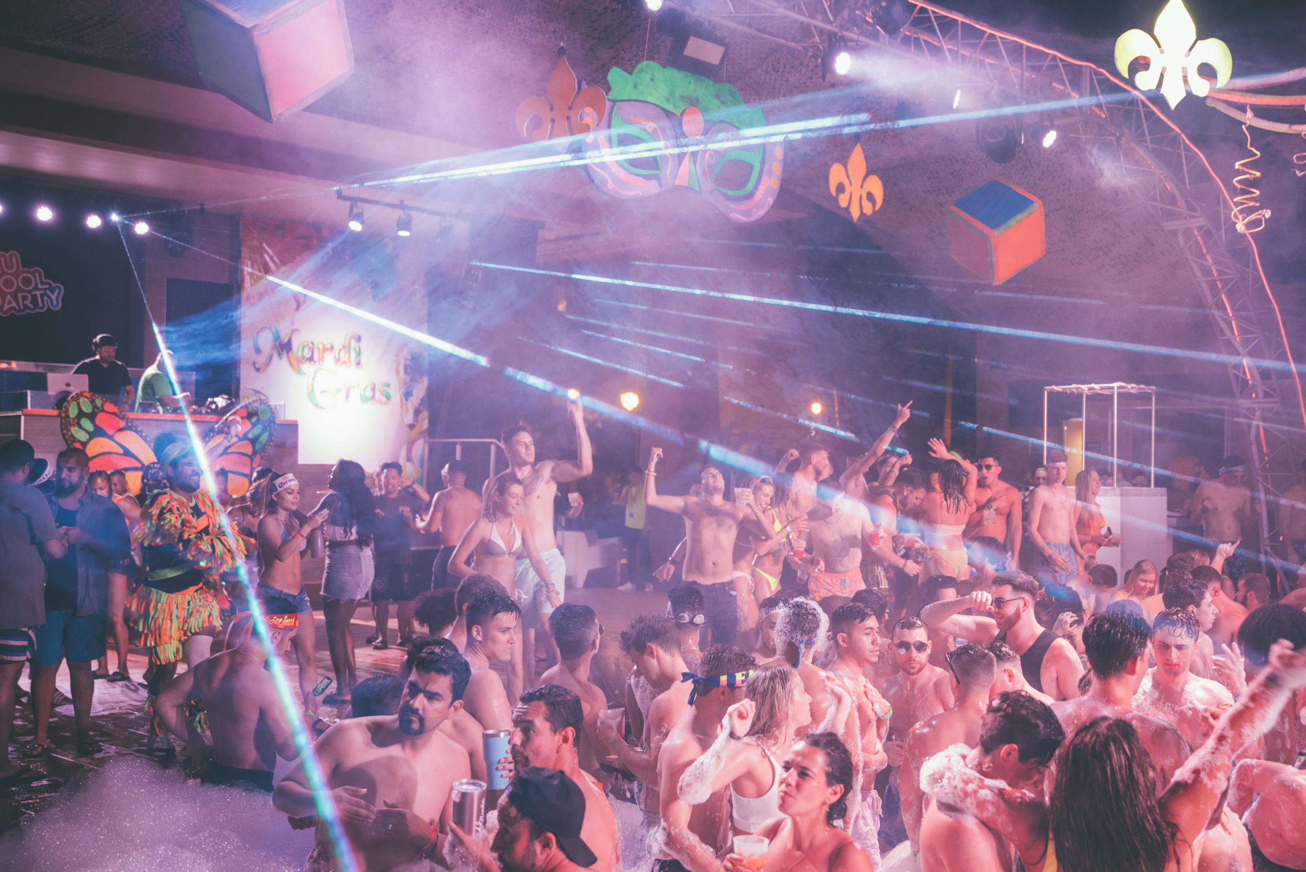 Lively indoor pool party featuring people enjoying a Mardi Gras themed celebration with foam and laser lights.
